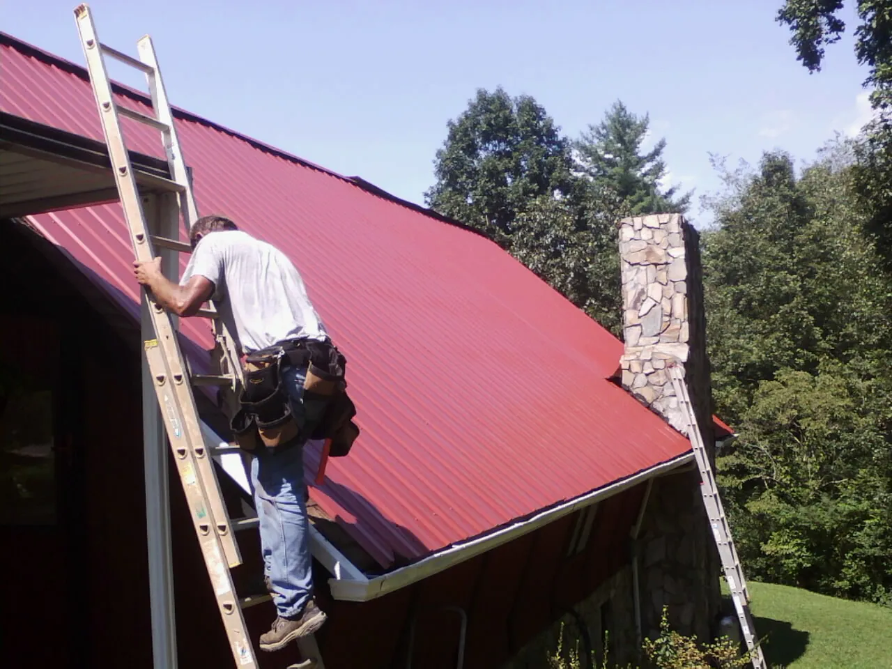 Roofer on a ladder beside a red metal roof and stone chimney — Soffit & Fascia Repair by Roofing Brothers in Patterson